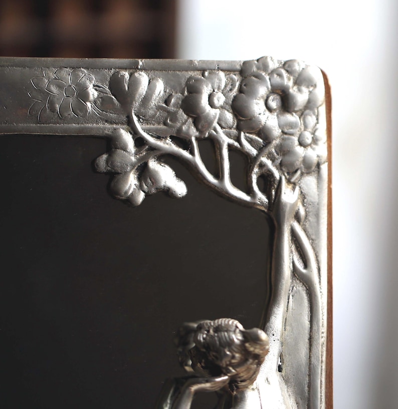 May include: A silver-toned picture frame with an ornate floral design. The frame is made of metal and has a mirror inside. The frame is in the Art Nouveau style.