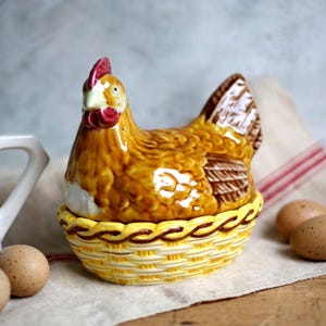 Puede incluir: Un portahuevos de cerámica con forma de gallina con base de cesta amarilla. La gallina es marrón y canela con cresta y barbilla rojas. Varios huevos marrones están junto al soporte, y una jarra blanca con cuchillos en el fondo.