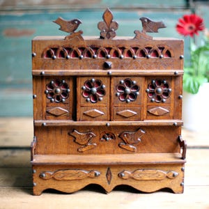 May include: A detailed wooden jewellery box with a rich, brown hue. It showcases floral and bird designs, a fleur-de-lis, and a red fabric interior. The box has a small bench-like base and is adorned with metal studs.