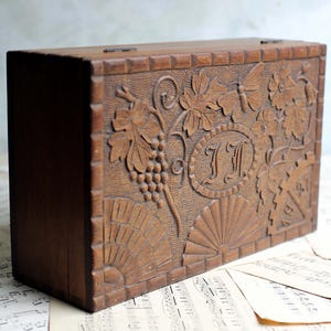 May include: A rectangular wooden box with carved grape vines, leaves, and a central monogram. The box is a warm brown color and has a hinged lid. The design includes a fan-like pattern at the bottom.