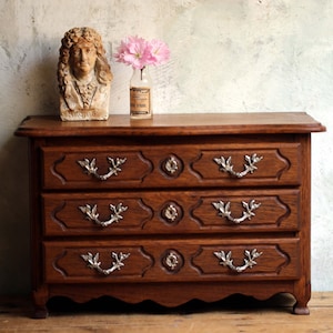 May include: A three-drawer wooden chest with ornate carvings and silver handles. The chest is made of dark wood and has a slightly curved bottom edge. A bust of a man with curly hair and a white collar sits on top of the chest. A small glass vase with pink flowers sits next to the bust.
