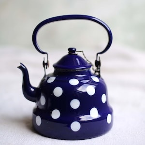 Puede incluir: Una tetera de esmalte azul marino con un estampado de lunares blancos. La tetera tiene un asa curva, un pico y una tapa con una pequeña cadena. Un artículo de cocina clásico.