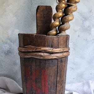 Op de afbeelding: Een hoge, cilindervormige houten emmer met verticale duigen, bijeengehouden door gedraaide, wijnrankachtige banden. Het donkere hout is voorzien van een rood geschilderd Romeins cijfer XI. Binnenin staan drie gedraaide, goudkleurige houten staven.