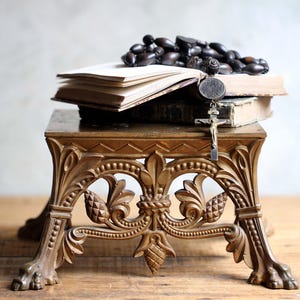 Puede incluir: Un soporte dorado ornamentado con un diseño floral detallado, que sostiene libros abiertos y un rosario. Un crucifijo cuelga de los libros. El soporte tiene patas decorativas y una parte superior detallada, creando una estética vintage.