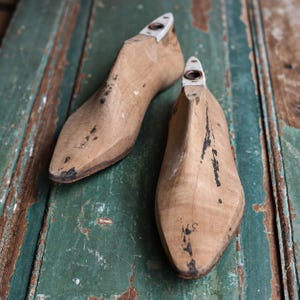 May include: A pair of light brown wooden shoe lasts with visible dark scuff marks and wear. Each form features a pointed toe, a contoured instep, and a white metal heel insert with a central circular opening. The wood grain is natural and textured.