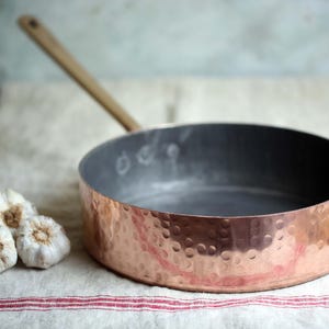 Può includere: Una padella color rame con finitura martellata e manico in legno. La padella è appoggiata su un panno bianco a righe rosse, accanto a tre teste d'aglio. La padella ha un interno scuro ed è progettata per cucinare.
