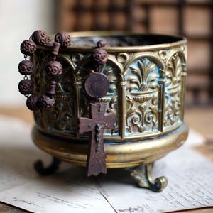 May include: An antique brass planter with ornate relief detailing and a dark brown rosary. The planter has a round base and three decorative feet. A wooden cross hangs from the rosary. The planter is on a piece of paper.