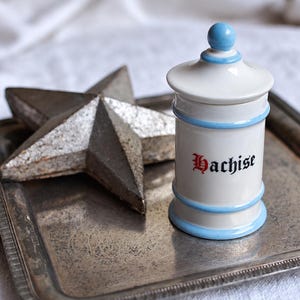 May include: A white ceramic apothecary jar with a lid, featuring light blue horizontal bands and the word "Hachise" printed in black gothic lettering. The jar is accompanied by a five-pointed star with a textured, metallic silver finish.
