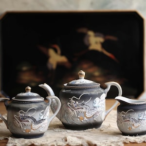 Op de afbeelding: Een vintage theeset met een zwart-wit drakenontwerp. De set bevat een theepot, een melkkan en een suikerpot. De achtergrond heeft een zwart dienblad met een schilderij van vogels.