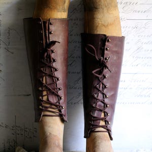 May include: A pair of antique brown leather boots with wooden forms inside. The boots have a lace-up design with brass eyelets and are displayed against a vintage background with faded script.