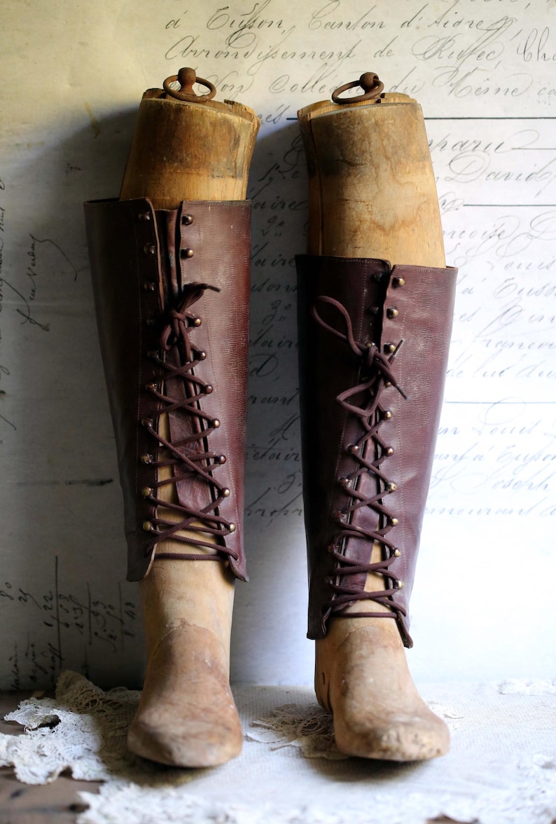 May include: A pair of antique brown leather boots with wooden forms inside. The boots have a lace-up design with brass eyelets and are displayed against a vintage background with faded script.