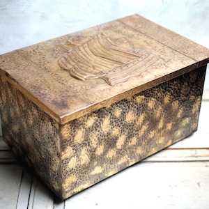 May include: A rectangular, antique-style box with a textured, mottled surface in shades of gold and brown. The lid features a raised relief of a sailing ship. The box has a hinged lid and is likely made of metal.