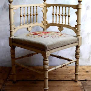 Peut inclure: Chaise d'angle crème avec un siège brodé de fleurs. Le dossier décoratif comporte des balustres verticaux et des accents dorés. Les pieds sont tournés et reliés par des entretoises, et le siège est recouvert d'un tissu vert clair avec un motif floral.