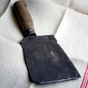 Op de afbeelding: Een vintage slagermes met een houten handvat en een zwart metalen blad. Het mes ligt op een witte linnen tafelkleed.