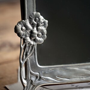 May include: A silver-toned picture frame with an ornate floral design. The frame has a rectangular shape and a beveled edge. The floral design is on the top and bottom of the frame. The frame is sitting on a piece of paper.