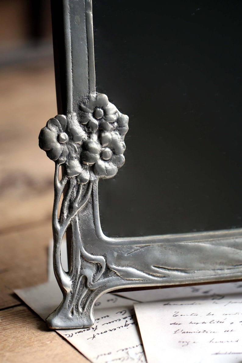 May include: A silver-toned picture frame with an ornate floral design. The frame has a rectangular shape and a beveled edge. The floral design is on the top and bottom of the frame. The frame is sitting on a piece of paper.