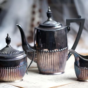 Puede incluir: Un juego de té plateado con tetera, azucarero y jarrita para la leche. El juego tiene un diseño acanalado en la parte inferior de cada pieza. La tetera tiene un pico largo y un asa negra.