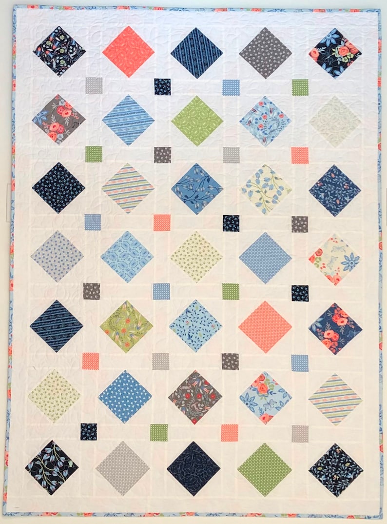 A quilt with a white background and a diamond pattern. The diamonds are in various colors and patterns, including blue, green, gray, pink, and floral. The quilt is edged with a light blue trim.