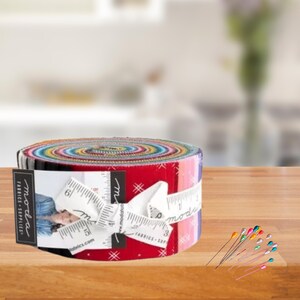 May include: A Moda Fabrics roll of 2.5 inch fabric strips with a red and white patterned design. The roll is tied with a white ribbon and a measuring tape. The Moda Fabrics logo is visible on the label.