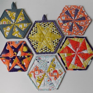 Könnte beinhalten: Sechs sechseckige Topflappen aus Stoff in verschiedenen Farben und Mustern. Die Topflappen haben ein wirbelndes Design mit einem Stern in der Mitte. Die Farben umfassen Orange, Gelb, Blau, Grün und Weiß.