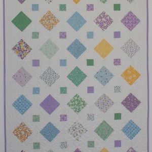 A white quilt with a purple border. The quilt is made up of a pattern of light purple, light blue, light green, and yellow diamond shapes with smaller squares of the same colors in between.