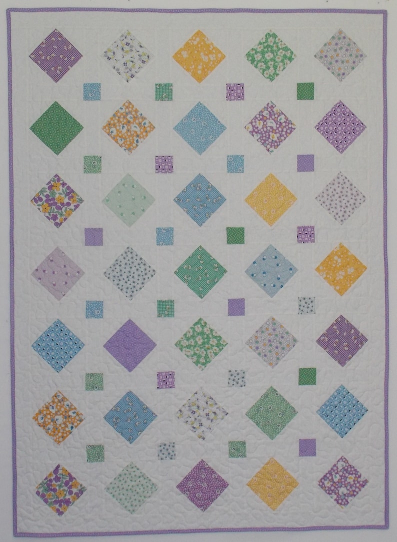 A white quilt with a purple border. The quilt is made up of a pattern of light purple, light blue, light green, and yellow diamond shapes with smaller squares of the same colors in between.