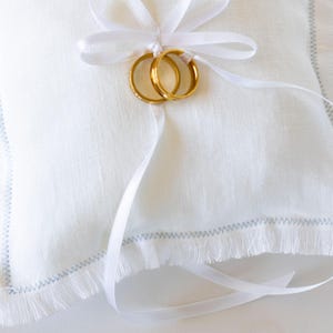 K&ouml;nnte beinhalten: Wei&szlig;es Leinen-Ringkissen mit ausgefransten Kanten und blauen N&auml;hten. Ein wei&szlig;es Satinband ist um das Kissen gebunden, mit zwei goldenen Eheringen.