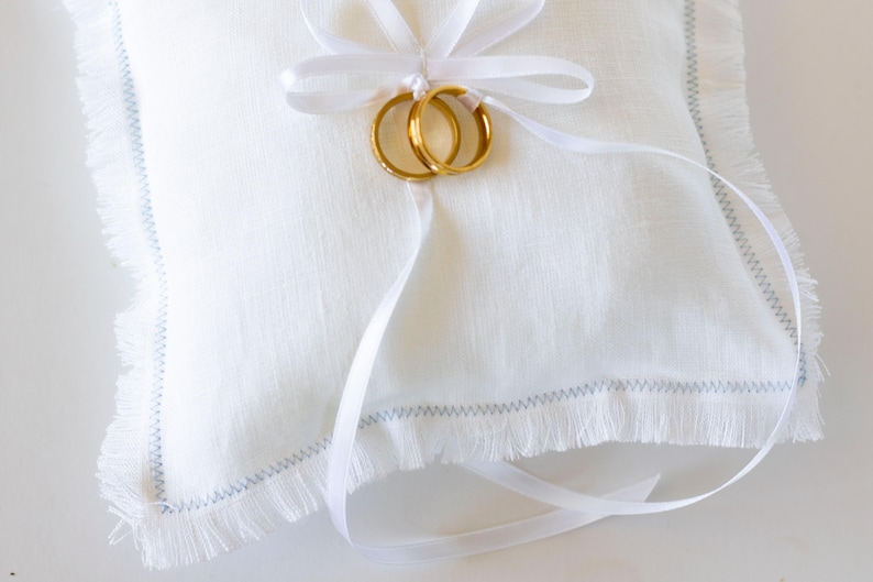 K&ouml;nnte beinhalten: Wei&szlig;es Leinen-Ringkissen mit ausgefransten Kanten und blauen N&auml;hten. Ein wei&szlig;es Satinband ist um das Kissen gebunden, mit zwei goldenen Eheringen.