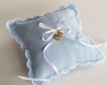 May include: A light blue ring pillow with white scalloped edges and a white satin ribbon tied in a bow. Two gold wedding bands are placed on top of the bow.