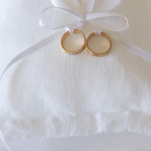 K&ouml;nnte beinhalten: Zwei goldene Eheringe auf einem wei&szlig;en Kissen mit einem wei&szlig;en Satinband.