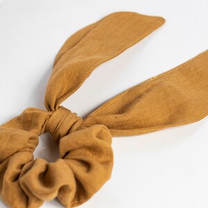 Könnte beinhalten: Ein braunes Stoffscrunchie mit zwei langen Stoffbändern. Das Scrunchie ist aus einem weichen, strukturierten Stoff.