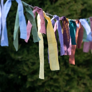 May include: A string of colorful fabric ribbons tied together and hanging from a branch. The ribbons are in shades of blue, green, yellow, purple, and brown.