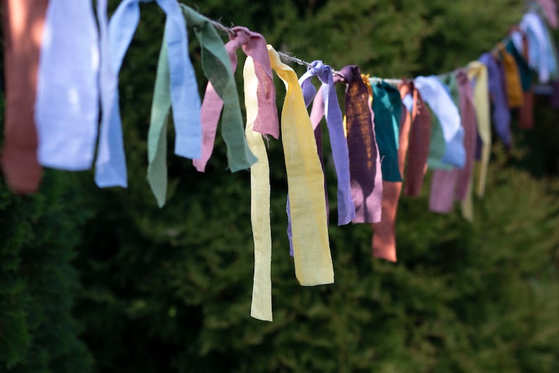 May include: A string of colorful fabric ribbons tied together and hanging from a branch. The ribbons are in shades of blue, green, yellow, purple, and brown.