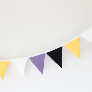 Könnte beinhalten: Eine Wimpelkette mit abwechselnden Farben in Schwarz, Gelb, Weiß und Lila. Die Kette ist an einer weißen Schnur befestigt und hängt an einer weißen Wand.