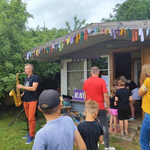 May include: A man plays a gold saxophone in front of a small building with a purple sign that says "KAV". The building is decorated with colorful flags. There are people standing in line to enter the building.