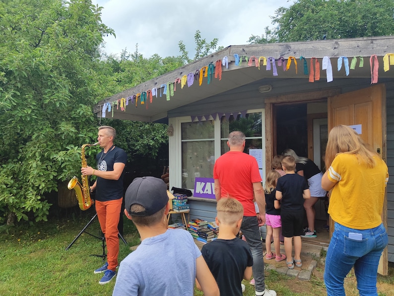 May include: A man plays a gold saxophone in front of a small building with a purple sign that says "KAV". The building is decorated with colorful flags. There are people standing in line to enter the building.