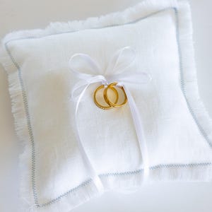 K&ouml;nnte beinhalten: Ein wei&szlig;es Ringkissen aus Leinen mit ausgefransten R&auml;ndern und einem hellblauen Besatz. Zwei goldene Eheringe liegen auf dem Kissen und sind mit einem wei&szlig;en Satinband gebunden.