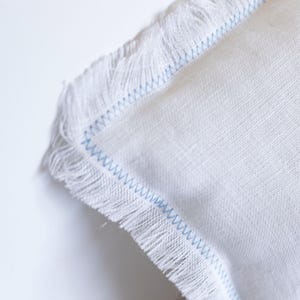 K&ouml;nnte beinhalten: Wei&szlig;es Kissen mit einem blauen, gesteppten Rand und ausgefransten Kanten.