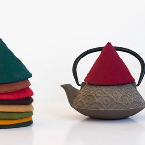 Peut inclure: Une pile de chapeaux de thé en feutre colorés, verts, bruns, rouges et jaunes. Une théière en fonte avec une anse noire et un chapeau de thé en feutre rouge se trouve à droite. La théière présente un motif décoratif.