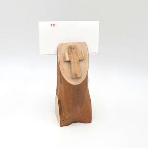 May include: A wooden cross sits atop a natural wood log, which is used as a card holder. The card holder has a blank card with the word "TO:" written on it.