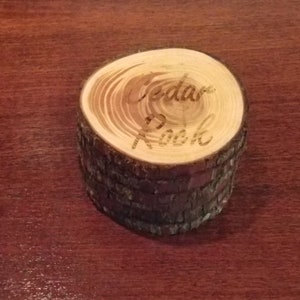 May include: A small wooden ring box made from a cedar log. The top of the box is a cross-section of the log with the words "Cedar Nook" engraved into the wood.