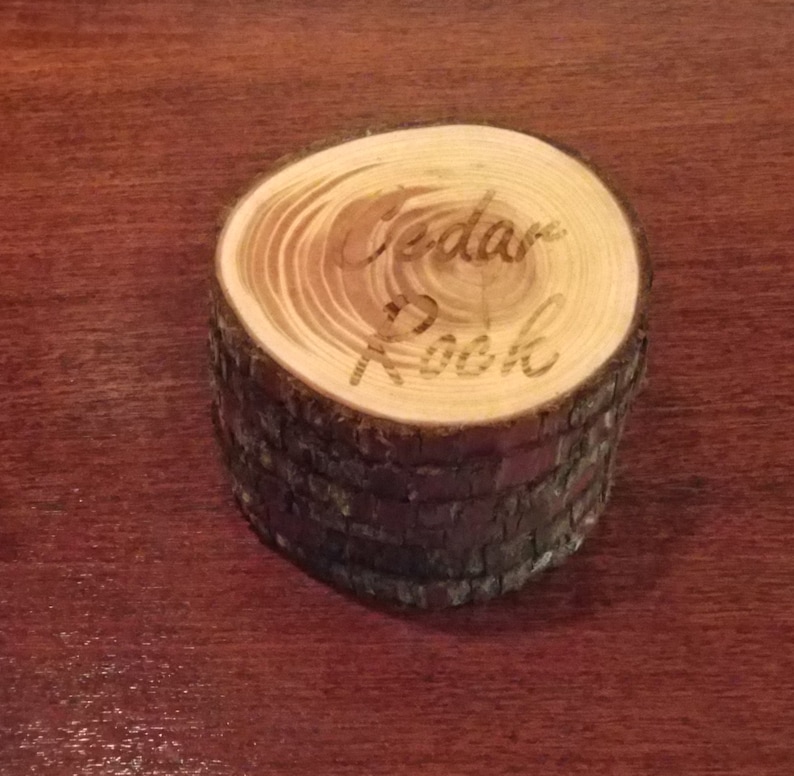 May include: A small wooden ring box made from a cedar log. The top of the box is a cross-section of the log with the words "Cedar Nook" engraved into the wood.