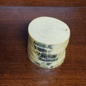May include: A stack of six round wooden coasters with a natural wood finish. The coasters have a dark brown wood grain pattern.