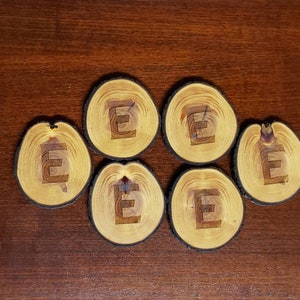May include: Six wooden coasters with a natural wood finish and a dark brown edge. Each coaster features a light brown letter "E" in the center.