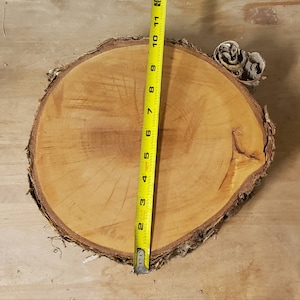 May include: A round, natural wood slice with a bark edge. A yellow measuring tape shows the diameter is approximately 10 inches. The wood has a light brown color with visible growth rings. Decorative wood slice for crafts and home decor.