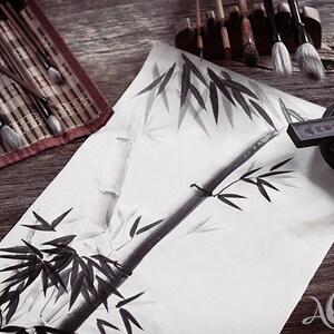 May include: An artistic still life featuring traditional calligraphy tools. The scene includes bamboo brushes, ink stones, a bamboo painting, and a red seal. The artwork depicts a bamboo stalk and leaves in black ink on white paper. The tools are arranged on a wooden surface.