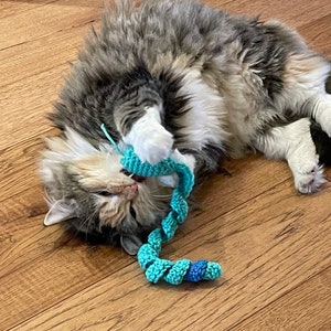 May include: A fluffy, multi-colored cat plays with a turquoise and blue crocheted cat toy on a wooden floor. The cat is lying on its back, paws playfully interacting with the toy.