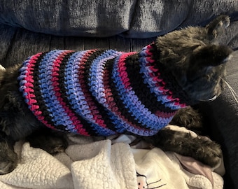 Könnte beinhalten: Eine schwarze Katze trägt einen farbenfrohen, gestreiften Häkelpullover. Der Pullover hat Bänder in Pink, Schwarz, Blau und Lila. Die Katze liegt auf einer weißen Decke mit einem Cartoon-Katzen-Gesicht.