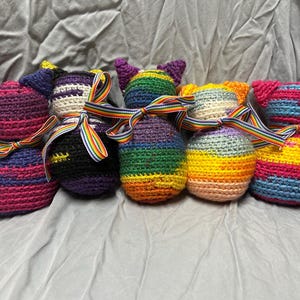 May include: Five handmade crochet cat-shaped plush toys arranged in a row. Each plush features a unique multicolored yarn pattern and is tied with a rainbow-striped ribbon around its neck. The cats have pointed ears and rounded bodies.
