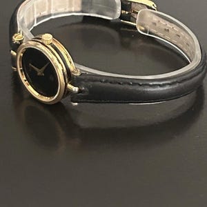 May include: A wristwatch featuring a round black dial with gold-tone hands and a gold-tone bezel. The watch has a thin black leather strap with a gold-tone buckle closure. The design includes a small crown on the side of the case for time adjustment.
